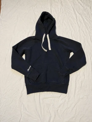 Mörkblå hoodie med vita snören - Mörkblå hoodie med stora fickor framtill och vita dragsnören i huvan. Klassisk passform och ribbade muddar vid ärmar och nederkant. Tillverkad i mjukt bomullsmaterial, perfekt för en avslappnad stil.