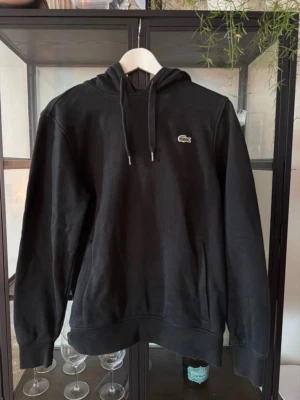 Svart hoodie från Lacoste Sport - Snygg svart hoodie från Lacoste Sport med klassisk krokodil-logga på bröstet. Hoodien har dragsko i huvan och en stor magficka. Perfekt för en chill och sportig stil. Materialet är mjukt och bekvämt, passar till det mesta.