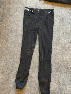 Svarta sportleggings med silikon - Säljer ett par svarta ridsportleggings från CRW med silikonprint på insidan av benen för extra grepp. De har bred resår i midjan med logga och dubbla knappar framtill. Materialet är stretchigt och följsamt, perfekt för träning eller ridning.