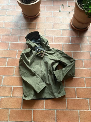 Olivgrön Arc'teryx Gore-Tex regnjacka - Snygg olivgrön regnjacka från Arc'teryx i slitstarkt Gore-Tex-material. Jackan har huva, hel dragkedja framtill och två sidofickor med dragkedja. Perfekt för blöta dagar och outdoor-äventyr. Minimalistisk design med logga på bröstet.