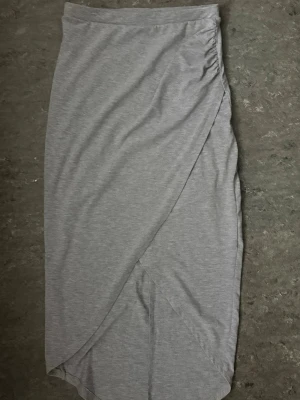 Grå omlottkjol från Gina Tricot - Snygg grå långkjol från Gina Tricot i mjukt bomullsmaterial. Kjolen har omlottdesign och lätt rynkning i sidan för en cool och avslappnad look. Perfekt att styla med en crop top eller hoodie.