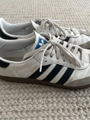 Adidas Samba vita och svarta sneakers - Klassiska Adidas Samba sneakers i vitt läder med svarta ränder och detaljer. Skorna har grå mocka framtill, bruna gummisulor och vita skosnören. Ikonisk blå Adidas-logga på plösen och gul SAMBA-tryck på sidan. Perfekta för en sportig och stilren look.