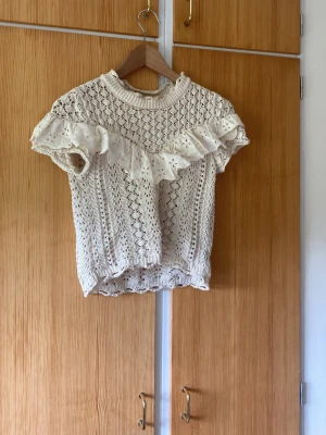 Vit stickad blus med volang - Supersöt vit stickad blus med korta ärmar och bred volangdetalj framtill. Blusen har ett fint hålmönster och ribbad kant vid hals och nederdel. Perfekt för dig som gillar romantisk och trendig stil.