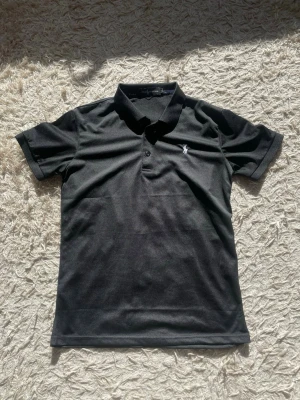 Svart Ralph Lauren Polo T-shirt - Helt oanvänd klassisk svart pikétröja från Polo Ralph Lauren med vit broderad logga på bröstet. Tröjan har krage, knappar framtill och är gjord i mjuk bomull. Perfekt till jeans eller shorts för en clean look.