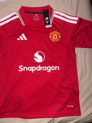 Manchester United matchtröja 26/27 B.Fernandes - Säljer en röd Manchester United matchtröja med B.Fernandes nummer 8 på ryggen från Adidas med klubbmärke på bröstet, vita detaljer och sponsorlogga 'Snapdragon' framtill. Tröjan har klassisk krage, vita ränder på axlarna och är tillverkad i lätt funktionsmaterial.