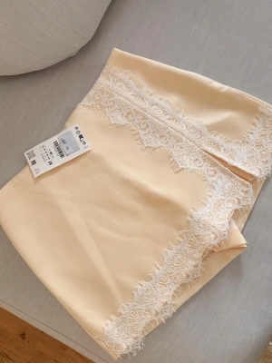 Beige kortkjol med spetsdetaljer - Supersnygg beige byxkjol med vita spetsdetaljer längs nederkant och framtill. Kjolen har en liten slits framtill och är gjord i ett mjukt, lätt material som känns lyxigt. Perfekt för dig som vill ha en feminin och trendig look.