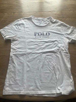 Vit t-shirt från Polo Ralph Lauren - Klassisk vit t-shirt från Polo Ralph Lauren med blå logga tryckt på bröstet. T-shirten har rund halsringning och korta ärmar. Tillverkad i mjuk bomull för en skön och avslappnad stil.
