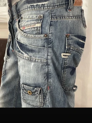 Diesel Blå cargobyxor i denim med fickor - Säljer ett par blå cargobyxor i denim med flera stora fickor och detaljer som knappar och nitar. Byxorna har en avslappnad passform och slitna tvättade partier för en cool streetwear-look. Perfekta för dig som gillar utility-stil och vill ha något unikt i garderoben. Jag är storlek xs för referens.