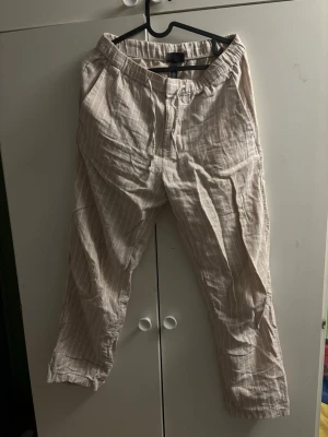 Beige linnebyxor med vita ränder - Säljer ett par beige linnebyxor med tunna vita ränder. Byxorna har avslappnad passform, resår i midjan och raka ben. Perfekta för en chill och luftig stil. Materialet är lätt och andas, vilket gör dem sköna att bära under varma dagar.