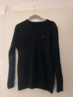 Svart stickad tröja med V-ringning - Svart stickad tröja från Ralph Lauren med V-ringning och lång ärm. Tröjan har ett diskret rött broderat logotyp på bröstet och är ribbstickad för en snygg passform. Perfekt för dig som gillar klassisk och stilren design. Använt bara en gång.  Har även samma i ljusrosa i storlek xs-s