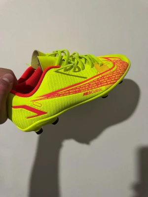 Neongula Nike Mercurial fotbollssko - Säljer en riktigt snygg Nike Mercurial fotbollssko i neongult med röda detaljer och text. Skon har låg profil, snörning och en ovandel i syntetmaterial för bra bollkontroll. Insidan är röd och yttersulan har fasta dobbar för spel på gräs.