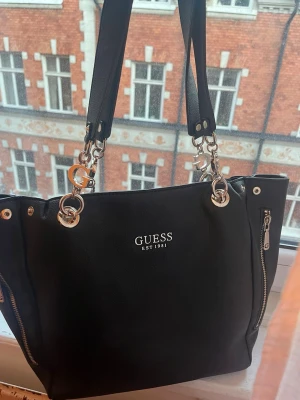 Svart handväska från Guess - Snygg svart handväska från Guess. Väskan har två dragkedjor framtill, dubbla axelband och logga i vitt på framsidan. Tillverkad i konstläder med stilrena metalldetaljer och G-hängen.