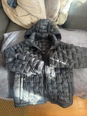 Svart pufferjacka med huva Everest - Snygg svart pufferjacka med huva och dragkedja framtill. Jackan har quiltad design och två sidofickor. Perfekt för kalla dagar och ger en modern look. Materialet är glansigt och vadderat för extra värme. köpt förra året och sparsamt använd. 