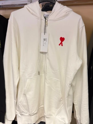 Vit hoodie med röd hjärtlogga - Snygg vit hoodie med dragkedja och huva. På bröstet finns en liten röd hjärtformad logga med bokstaven A. Hoodien har känguruficka framtill och ribbade muddar vid ärmar och nederkant. Perfekt för en avslappnad och trendig stil.