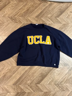 Mörkblå UCLA Russell sweatshirt  - Mörkblå sweatshirt från Russell Athletic i storlek XL med stor gul UCLA-text på bröstet. Klassisk rund halsringning och mjukt material, perfekt för en avslappnad och sportig stil.