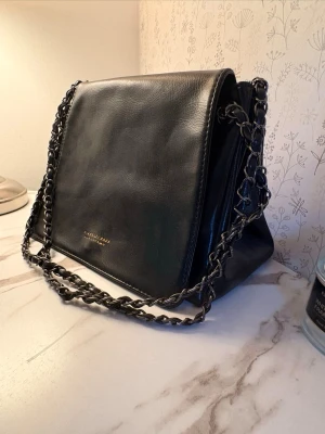 Svart axelväska med kedjerem - Snygg svart axelväska från Shein med stilren design och kedjerem i metall. Väskan är gjord i glansigt konstläder och har ett diskret guldfärgat logotryck på framsidan. Perfekt storlek för det viktigaste och ger en trendig touch till din outfit. Knappt använd 5 gånger. Har en till som liknar denna. 