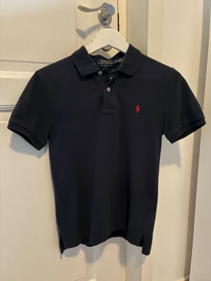 Mörkblå piké Ralph Lauren - Klassisk mörkblå pikétröja från Polo Ralph Lauren. Tröjan har två knappar vid halsen och den ikoniska röda logotypen broderad på bröstet. Perfekt till jeans eller shorts för en clean look. Storleken är junior L (14-16). Inköpt på Kidsbrandstore. 