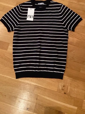 Navyvit randig t-shirt från Zara - Svart t-shirt från Zara med vita horisontella ränder och klassisk rund hals. Modellen har korta ärmar och ribbade kanter. Tillverkad i mjuk bomull som känns skön mot huden. Enkel och stilren design som funkar till det mesta.