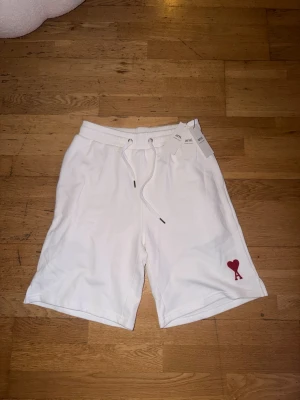 Vita shorts från ami - Snygga vita shorts med snörning i midjan från ami och en liten röd hjärtdetalj med bokstaven A på vänster ben. Mjukt material och relaxed fit, perfekta för sommaren.