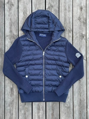 La Santino Quiltad Cardigan med Luva – Marinblå | Strl XS - Stilren marinblå La Santino cardigan med quiltad framsida och huva. Kombinationen av den vadderade framsidan och de stickade ärmarna ger både värme och en exklusiv look. Passar perfekt till både vardag och finare tillfällen.  ✔ Mycket fint skick ✔ Storlek Xs ✔ Marinblå färg ✔ Quiltad framsida med huva ✔ Stickade ärmar och rygg ✔ Mjuk och bekväm kvalitet Hör gärna av dig om du har några frågor eller vill ha fler bilder. Jag är alltid öppen för bud! 😊