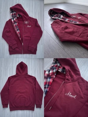 Vinröd hoodie med rutigt foder - Säljer en vinröd hoodie med dragkedja och huva. Insidan av huvan har ett rutigt foder i rött, vitt och blått. På bröstet finns en broderad text 'Marcell' i vitt. Hoodien har ribbade muddar och fickor framtill.