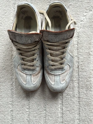 Maison Margiela ”Broken mirror” - Unika sneakers från Maison Margiela (BROKEN MIRROR) i metallic silverfärgat läder med beige snörning och bruna detaljer. Klassisk low-top siluett och gummisula. Märkets ikoniska etikett på plösen och vintageinspirerad design. Perfekta för dig som gillar statement skor. 