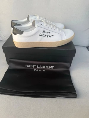 Saint Laurent vita sneakers med svart detalj - Snygga vita sneakers från Saint Laurent med svart hälparti och handskriven logga på sidan. Skorna har en beige sula och är tillverkade i läder med klassisk låg profil. Perfekta för en stilren och trendig look.
