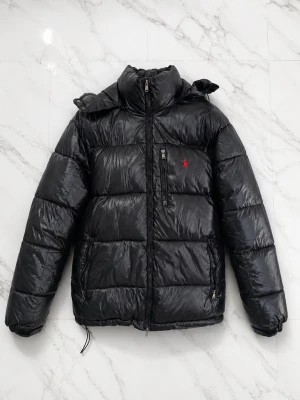 Jacka Polo Ralph Lauren - Snygg svart dunjacka från Polo Ralph Lauren med hög krage och dragkedja framtill. Jackan har två sidofickor med dragkedja och den klassiska röda loggan broderad på bröstet. Materialet är glansigt och ger en modern look. Perfekt för kalla vinterdagar.