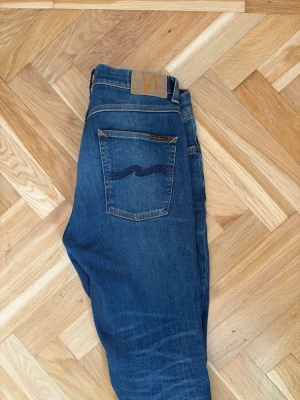 Blå jeans från Nudie Jeans - Säljer ett par klassiska blå jeans från Nudie Jeans med fem fickor och snygga slitningar framtill. Jeansen har en rak passform och är tillverkade i denim med en cool vågdetalj på bakfickan. Perfekta för en avslappnad och trendig look.