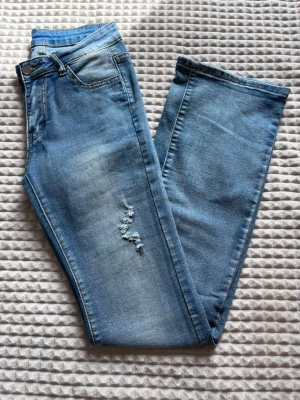 Ljusblå flare jeans med slitning - Säljer ett par ljusblå flare jeans och en snygg slitning på ena benet. Jeansen har klassisk femficksdesign, bälteshällor och stängs med knapp och dragkedja. Aldrig använt då jag helt glömde bort att jag har de. 