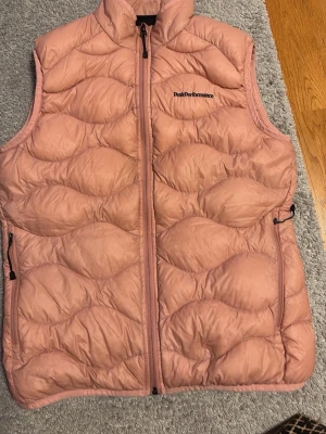 Rosa dunväst från Peak Performance - Säljer en snygg rosa dunväst från Peak Performance med vågigt quiltat mönster. Västen har hög krage, dragkedja framtill och två sidofickor med dragkedja. Perfekt för lager på lager och ger en cool look till din outfit.