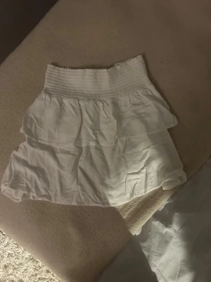 Vit volangkjol med smock - Söt vit kjol med bred smock i midjan och dubbla volanger. Kjolen har en luftig och lätt känsla, perfekt för varma dagar. Materialet känns mjukt och flowy, och modellen är kort och feminin. Använd fåtal gånger. 