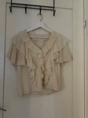 Beige blus med volanger och v-ringning - Supersöt beige blus med v-ringning och stora volanger framtill och på ärmarna. Blusen har en luftig och flowy passform, perfekt för en trendig och avslappnad look. Tillverkad i ett tunt, lätt material som ger en romantisk vibe.