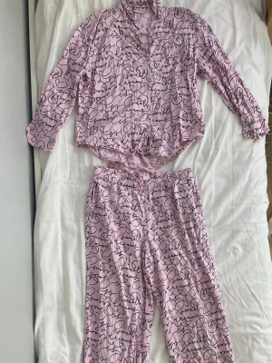 Rosa pyjamas med textmönster - Säljer en söt rosa pyjamas med svart textmönster där det står 'sweet dreams' och små hjärtan. Setet består av en långärmad skjorta med knappar och en bröstficka samt matchande byxor med resår i midjan. Perfekt för mysiga kvällar hemma. Obs: hål på baksidan