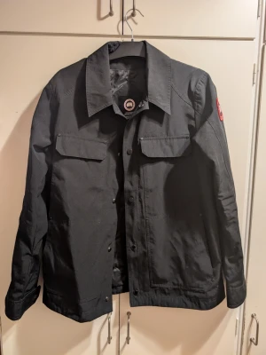 Canada Goose Vår/Höst - Stilren svart overshirt med två bröstfickor och knappstängning framtill. Bra för höst och vår, kan bäras under vintern med varmt underlag.