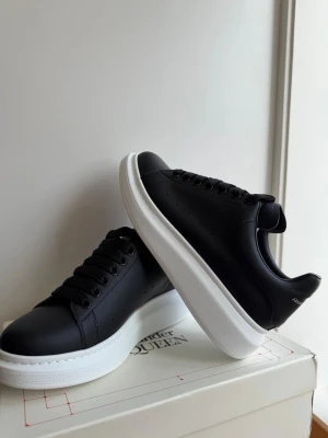 Svarta sneakers från Alexander McQueen - Använda några fåtal gånger, allt finns med! Stilrena svarta sneakers från Alexander McQueen med vit, chunky platt sula och klassisk snörning. Skorna är tillverkade i slätt skinn och har en minimalistisk design som passar perfekt till trendiga outfits.