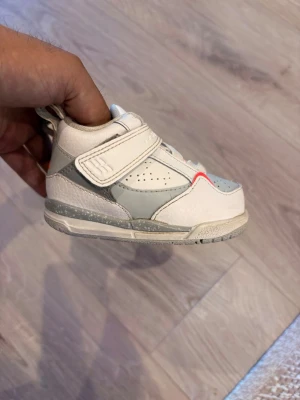 Jordan sneakers för barn, vit/grå - Snygga små Jordan sneakers i vitt och ljusgrått med orange detaljer. Skorna har kardborreband för enkel påtagning och är tillverkade i en kombination av läder och syntetmaterial. Perfekta för dig som vill ha stilrena och sportiga skor till de minsta.