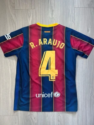 Barcelona matchtröja R. Araujo #4 - Säljer en officiell FC Barcelona matchtröja med Ronald Araujo och nummer 4 på ryggen. Tröjan har blå och vinröda vertikala ränder, gula detaljer och klubbmärke på bröstet. Tillverkad i lätt och ventilerande material, perfekt för fotboll eller träning. Mycket bra skick och är storlek M. Hör av dig vid frågor eller liknande!