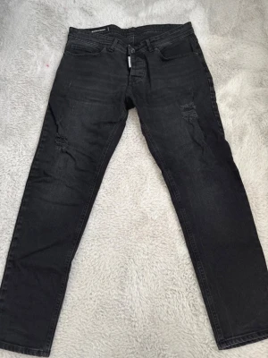 Svarta jeans  Dsquared2 - Säljer ett par svarta jeans från Dsquared2 med snygga slitningar på låren. Jeansen har klassisk femficksmodell, rak passform och detaljerad logotyp vid gylfen. Materialet är denim i bomull som ger en skön känsla och cool look. Dem är inte äkta.