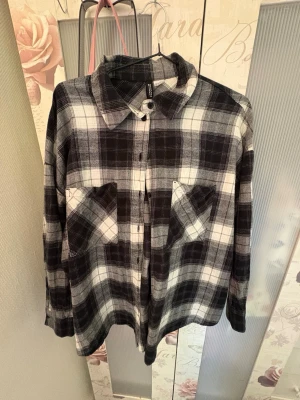 Rutig skjorta från H&M Divided - Säljer en svartvit rutig skjorta från H&M Divided i storlek S. Skjortan har klassisk krage, knäppning framtill och två bröstfickor. Perfekt för en avslappnad och trendig look. Tillverkad i mjuk bomull för skön känsla.