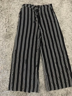 Svarta linnebyxor med vita ränder - Snygga svarta linnebyxor med vita vertikala ränder och bred resår i midjan. Byxorna har knytband i midjan och vida ben för en avslappnad och trendig look. Perfekta för dig som gillar en chill och stilren stil.