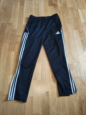 Svarta Adidas träningsbyxor herr - Säljer ett par svarta träningsbyxor från Adidas med klassiska vita ränder längs benen och logga på låret. Byxorna har resår i midjan, dragkedjor vid bensluten och är gjorda i ett mjukt, stretchigt material som passar perfekt till träning eller chill.