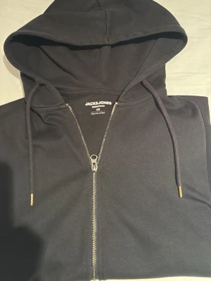 Svart zip hoodie från Jack & Jones - Säljer en svart hoodie från Jack & Jones Essentials med dragkedja framtill och justerbar huva med snören. Klassisk och enkel design, perfekt till vardags. Tillverkad i mjukt material för skön känsla. Metalländar på snörena och ribbade muddar vid ärmslut.