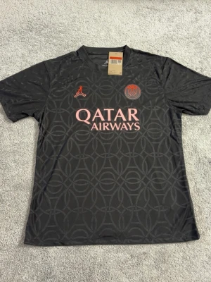 Svart PSG matchtröja med rosa tryck - Snygg svart matchtröja från PSG med diskret mönster och rosa detaljer. Framsidan har Qatar Airways-logga och PSG-emblem samt Jordan-logga. Perfekt för fotbollsträning eller att bära till vardags för att visa ditt lagstöd.