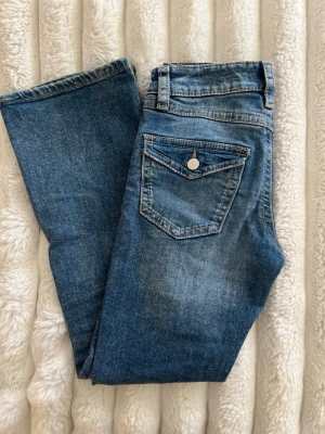 Blå bootcut jeans - Säljer ett par blå bootcut jeans med slitningar och hål på knäna. Jeansen har fem fickor, inklusive en bakficka med lock och knapp. Klassisk denim med ljus tvätt och råa detaljer för en trendig look.