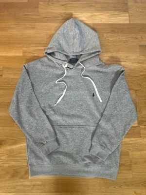Grå hoodie från Polo Ralph Lauren - Snygg grå hoodie från Polo Ralph Lauren med klassisk huva och vita dragsnören. Har en broderad marinblå logga på bröstet och en stor magficka. Tillverkad i mjukt sweatshirtmaterial och har ribbade muddar vid ärmar och midja. Perfekt för en avslappnad stil.