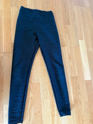 Svarta leggings med vågigt mönster - Säljer ett par svarta leggings med vågigt, strukturerat mönster. De har hög midja och är stretchiga för en bekväm passform. Perfekta till en avslappnad look eller när du vill ha något enkelt men ändå snyggt.