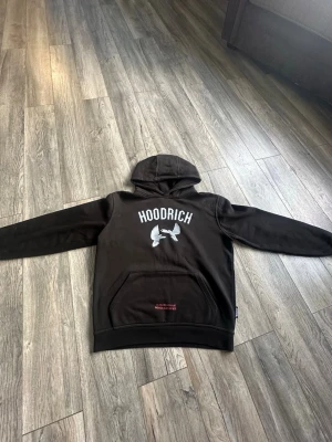 Svart hoodie med tryck från Hoodrich - Svart hoodie med stor huva och tryck fram och bak. På framsidan står det 'HOODRICH' med en vit duva under, och på baksidan finns två vita duvor samt röd text. Hoodien har magficka och ribbade muddar. Perfekt för en streetwear-look.