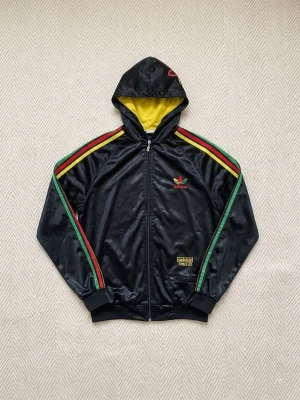 Adidas Chile - Snygg svart hoodie från Chile Adidas med dragkedja och huva. Fodret i huvan är gult och ärmarna har gröna, gula och röda ränder. Adidas-logga på bröstet och broderad detalj på huvan. Tillverkad i glansigt syntetmaterial.