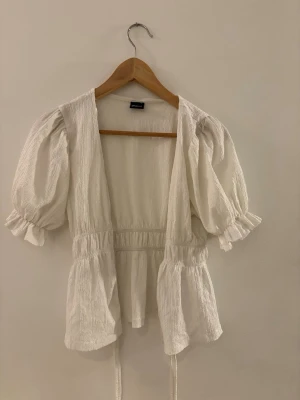 Vit puffärmad blus med knytband - Söt vit blus med puffiga korta ärmar och resår i midjan. Blusen har struktur i tyget och volangdetaljer vid ärmslut och nederkant. Knytband i midjan för en justerbar passform. Perfekt för en trendig och avslappnad look.